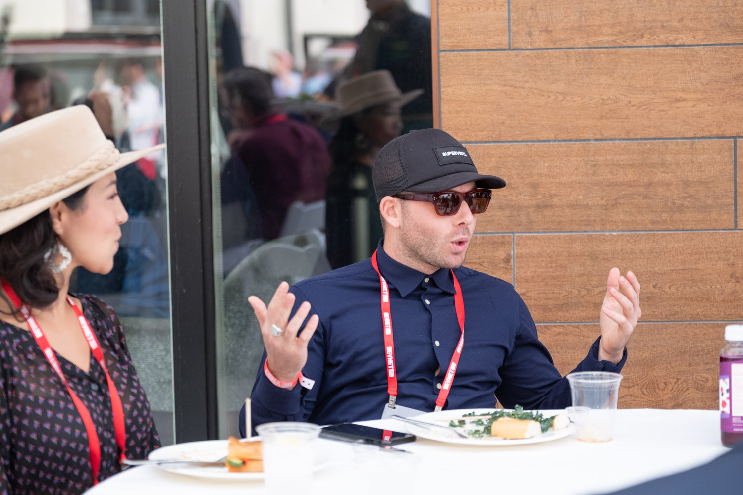 A man in sunglasses and a navy shirt gestures while talking at a table with a plate of food.