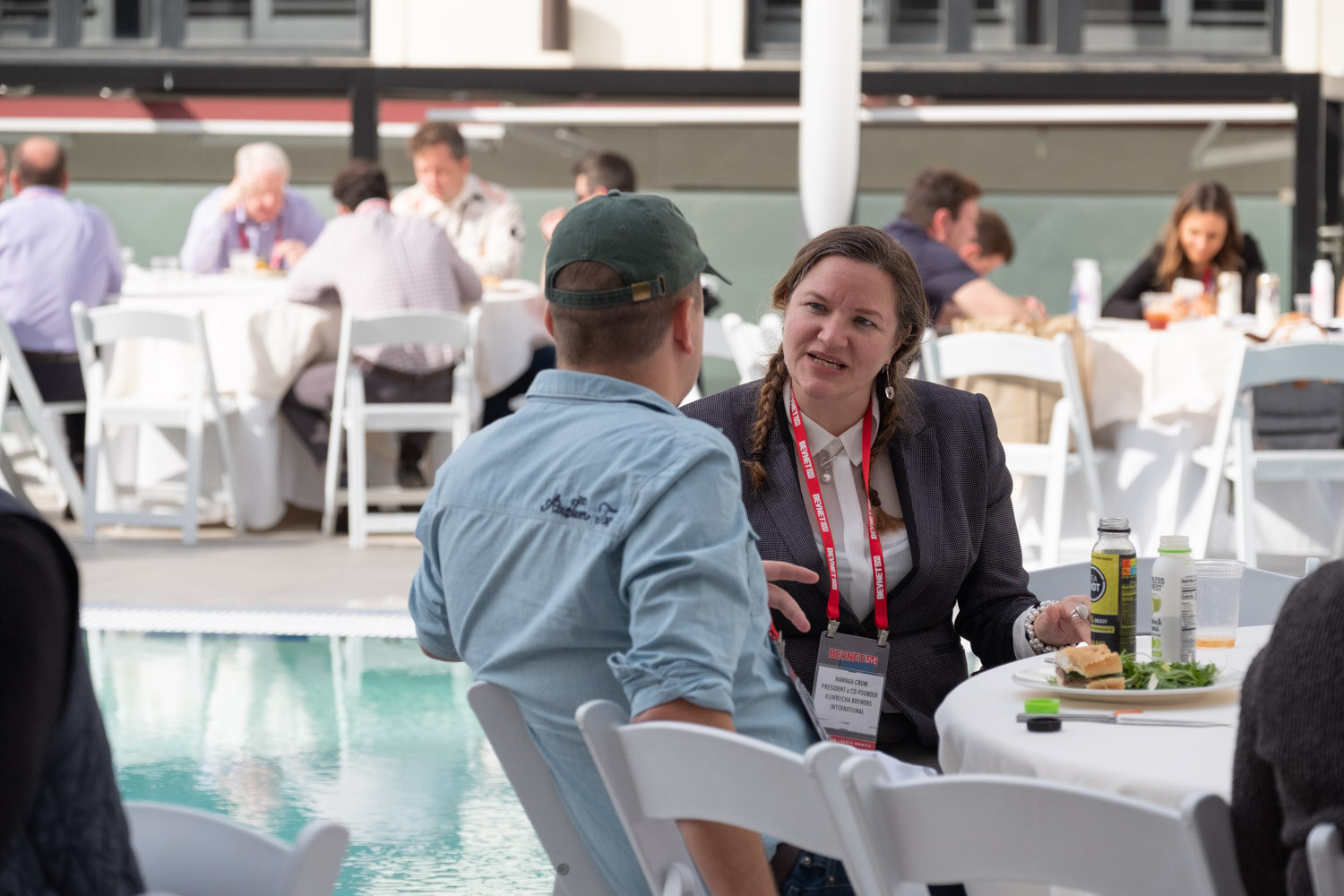 A man in a green cap and a woman in a dark blazer sit at a table by the pool with sandwiches and drinks.