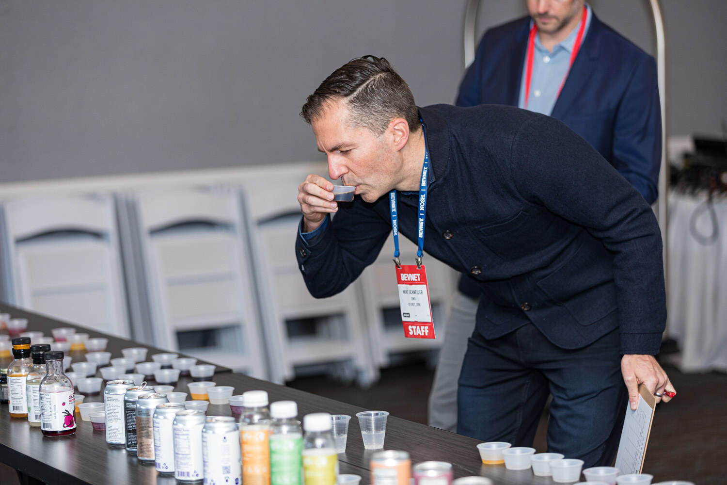 A man with a lanyard and a staff badge sips from a small cup at a table lined with bottles, cans and sample cups.
