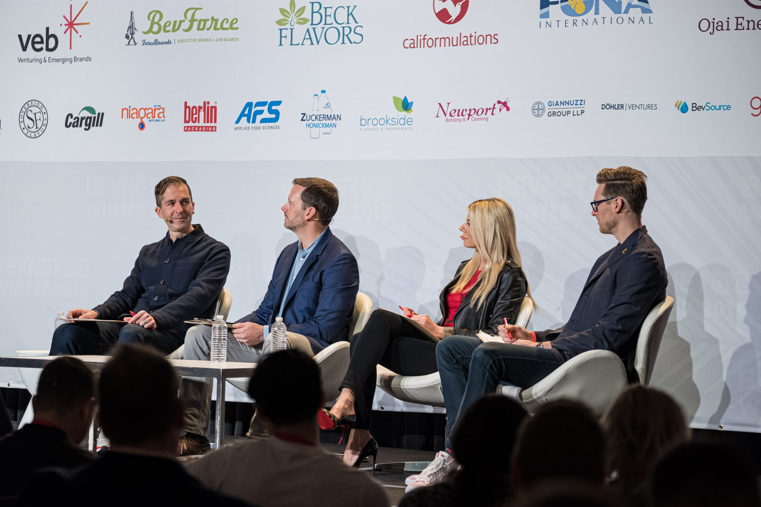 Four people with clipboards sit on white chairs on a stage in front of a sponsor wall with the audience in silhouette.