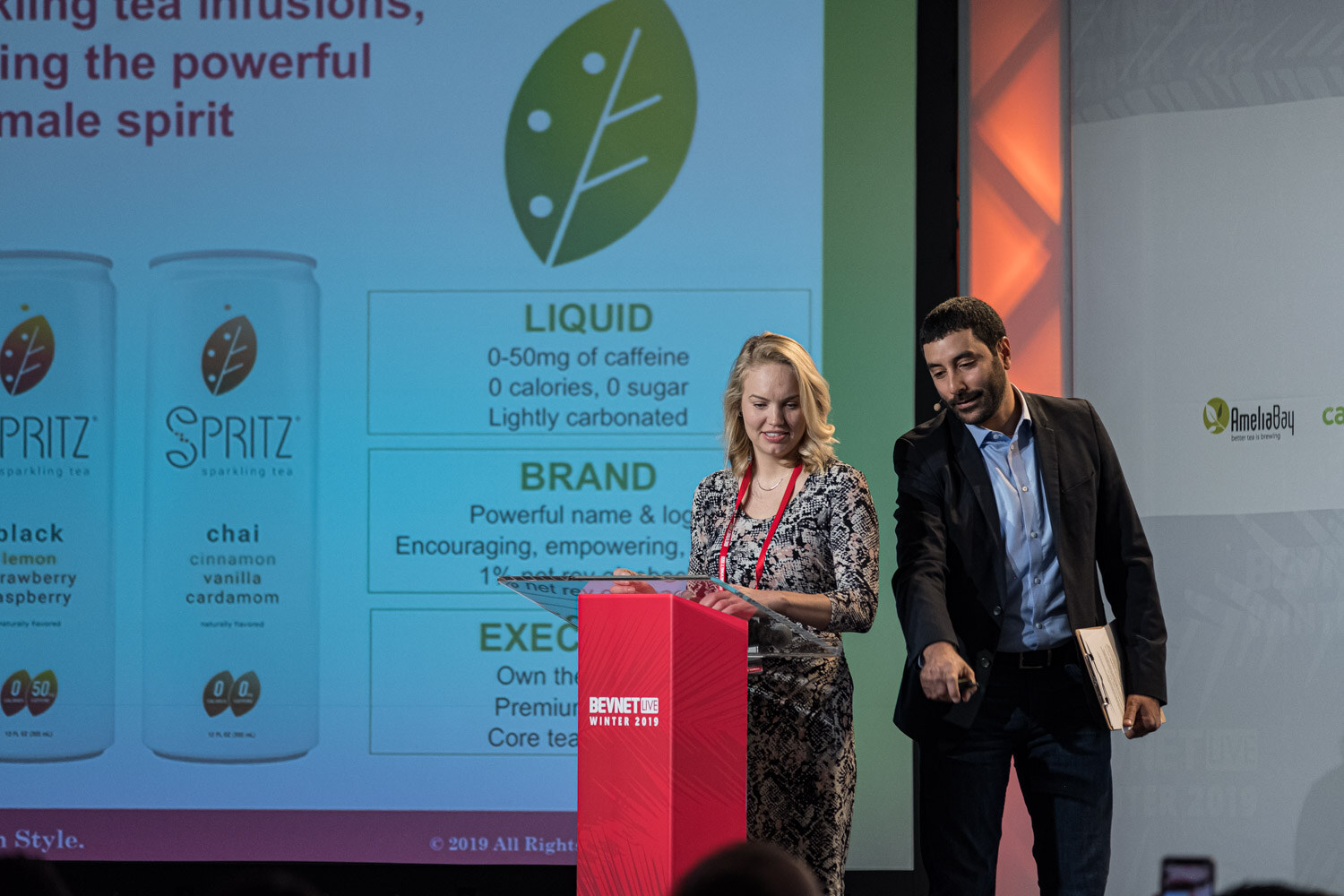A woman in a patterned dress stands at the lectern with a man in black beside her and a screen reading Spritz sparkling tea.