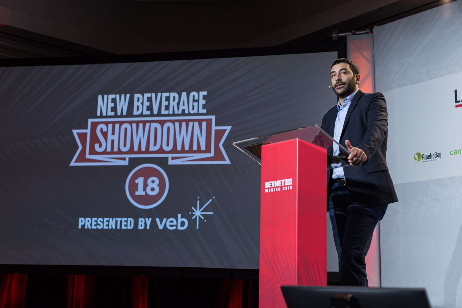 A man in a dark blazer speaks at a red lectern beside a screen reading New Beverage Showdown 18, presented by veb.