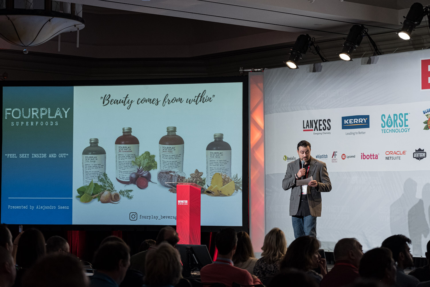 A man in a brown jacket stands by a lectern beside a screen showing bottles of juice and the name Fourplay Superfoods.