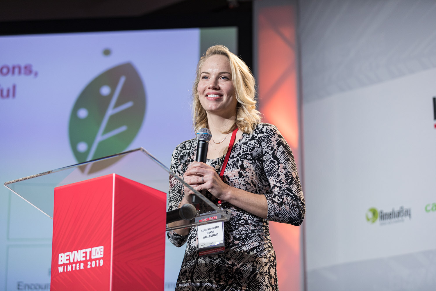 A blonde woman in a patterned dress with a lanyard smiles beside a lectern in front of a green Spritz sparkling tea slide.