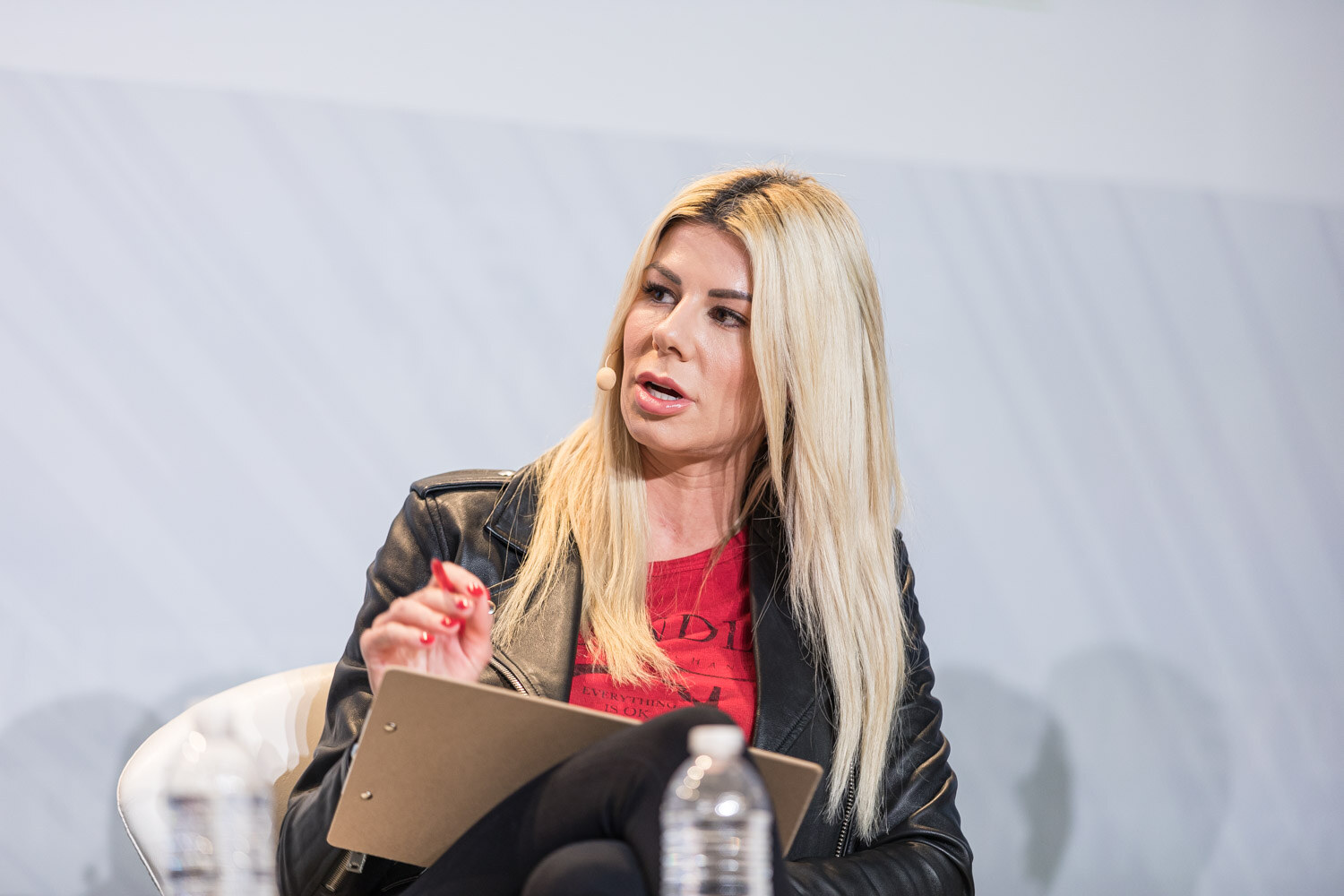 A woman in a black leather jacket and red top holds a clipboard and a pen while seated on a white chair.