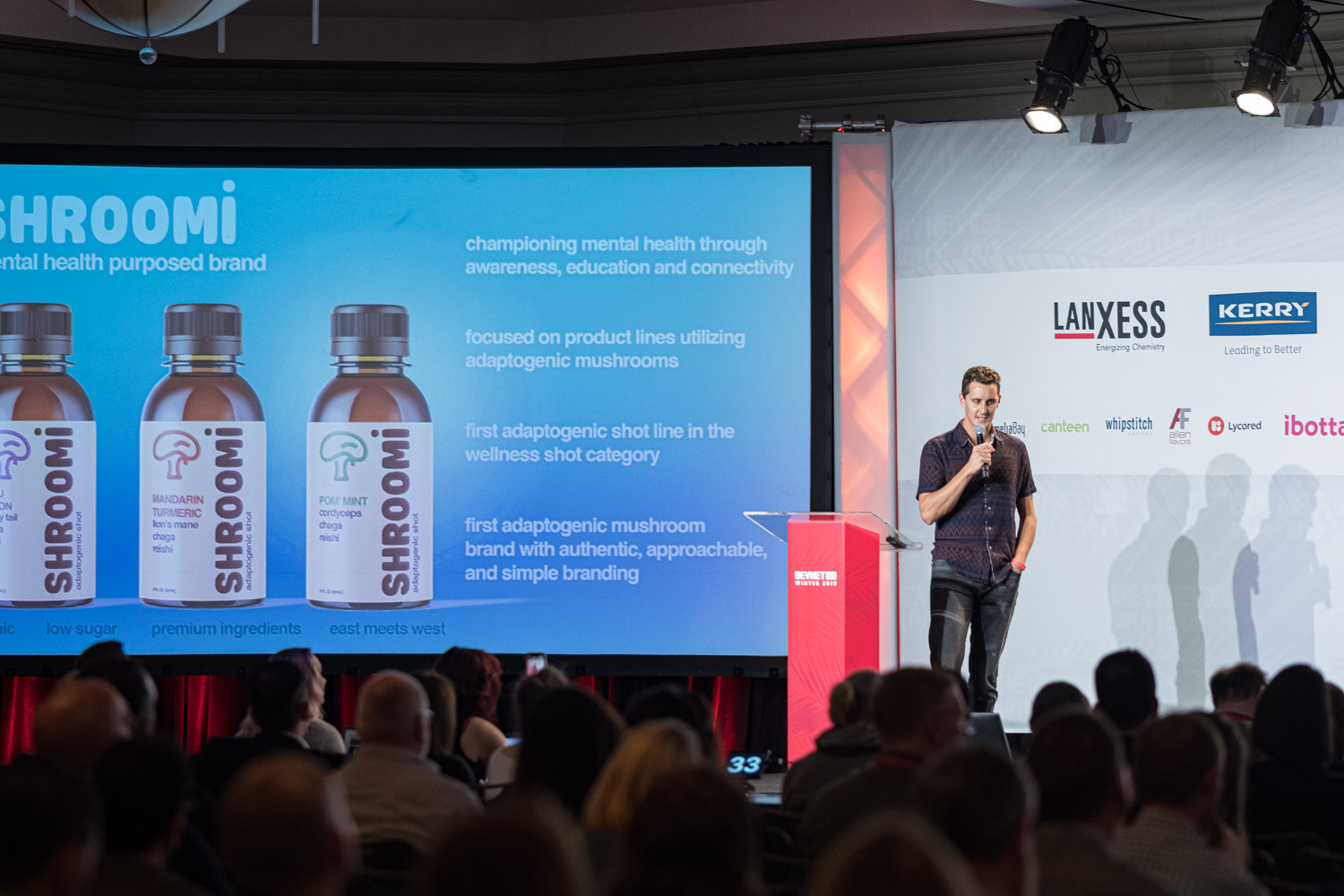 A man in a dark shirt stands at the red lectern beside a screen showing three Shroomi bottles and text about adaptogenic mushrooms.