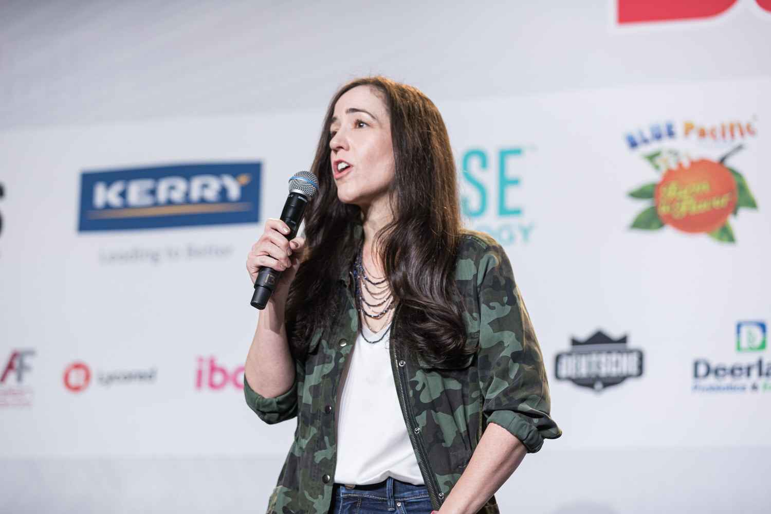 A woman with long dark hair and a camo jacket holds a microphone and gestures on stage in front of Kerry and Deerland logos.