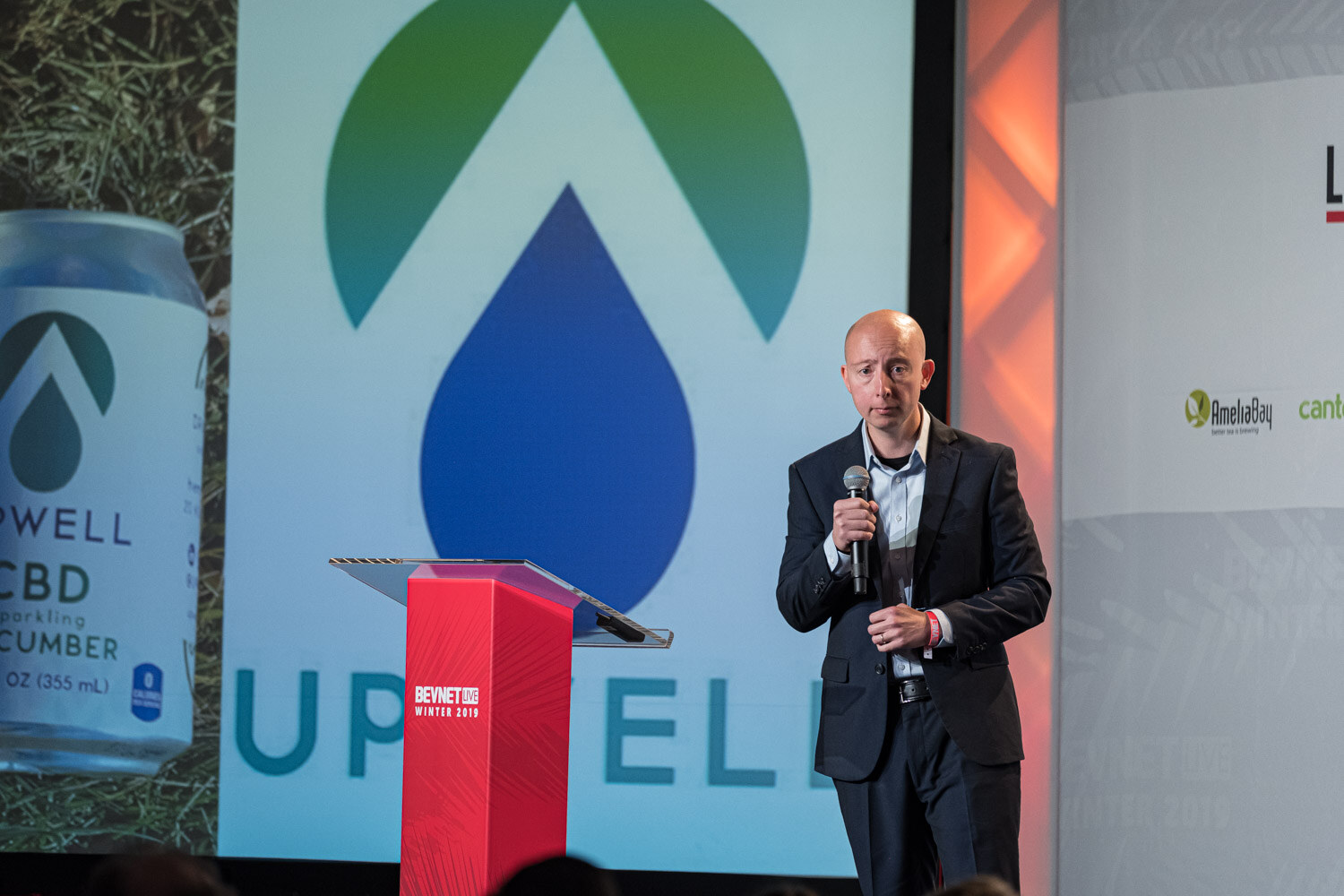 A man in a dark jacket holds a microphone at the lectern in front of a large screen showing an Upwell CBD cucumber water can.