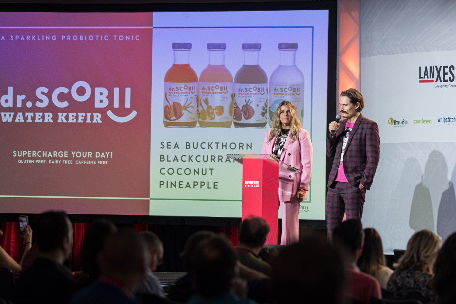A woman in a pink suit and a man in a plaid suit stand at the lectern in front of a screen reading dr.Scobii Water Kefir with flavors including sea buckthorn and coconut pineapple.