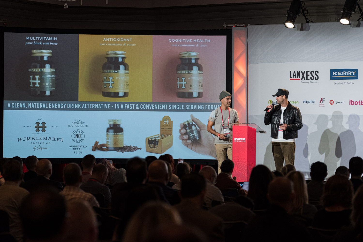 Two men stand at the lectern in front of a screen showing amber bottles and the text A clean, natural energy drink alternative with Humblemaker branding.