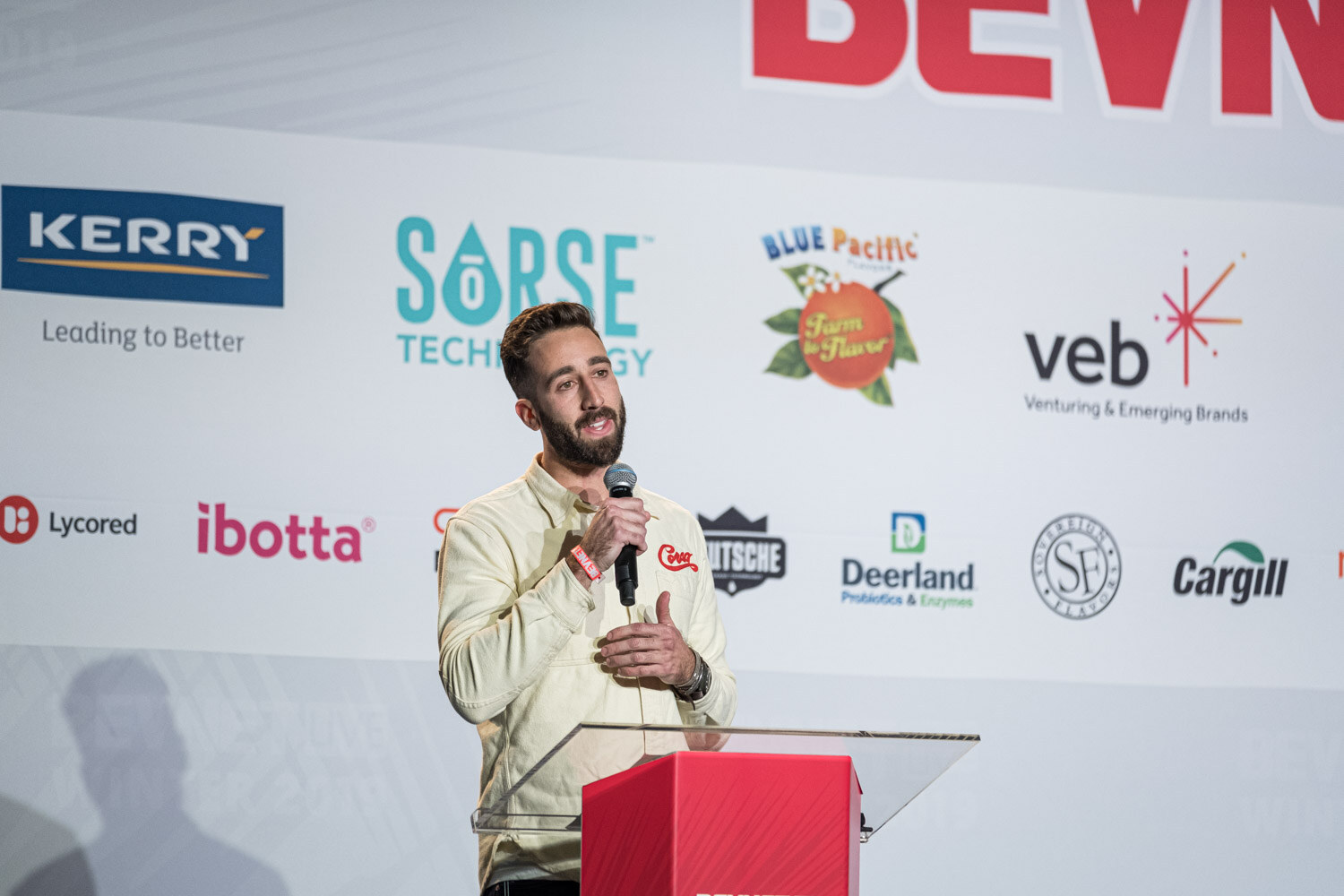 A bearded man in a cream jacket holds a microphone at a clear lectern in front of the sponsor wall.