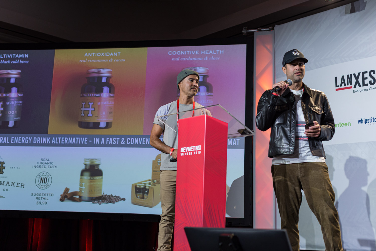 A man in a cap and a man in a leather jacket stand at the lectern in front of a screen showing amber bottles and boxes.