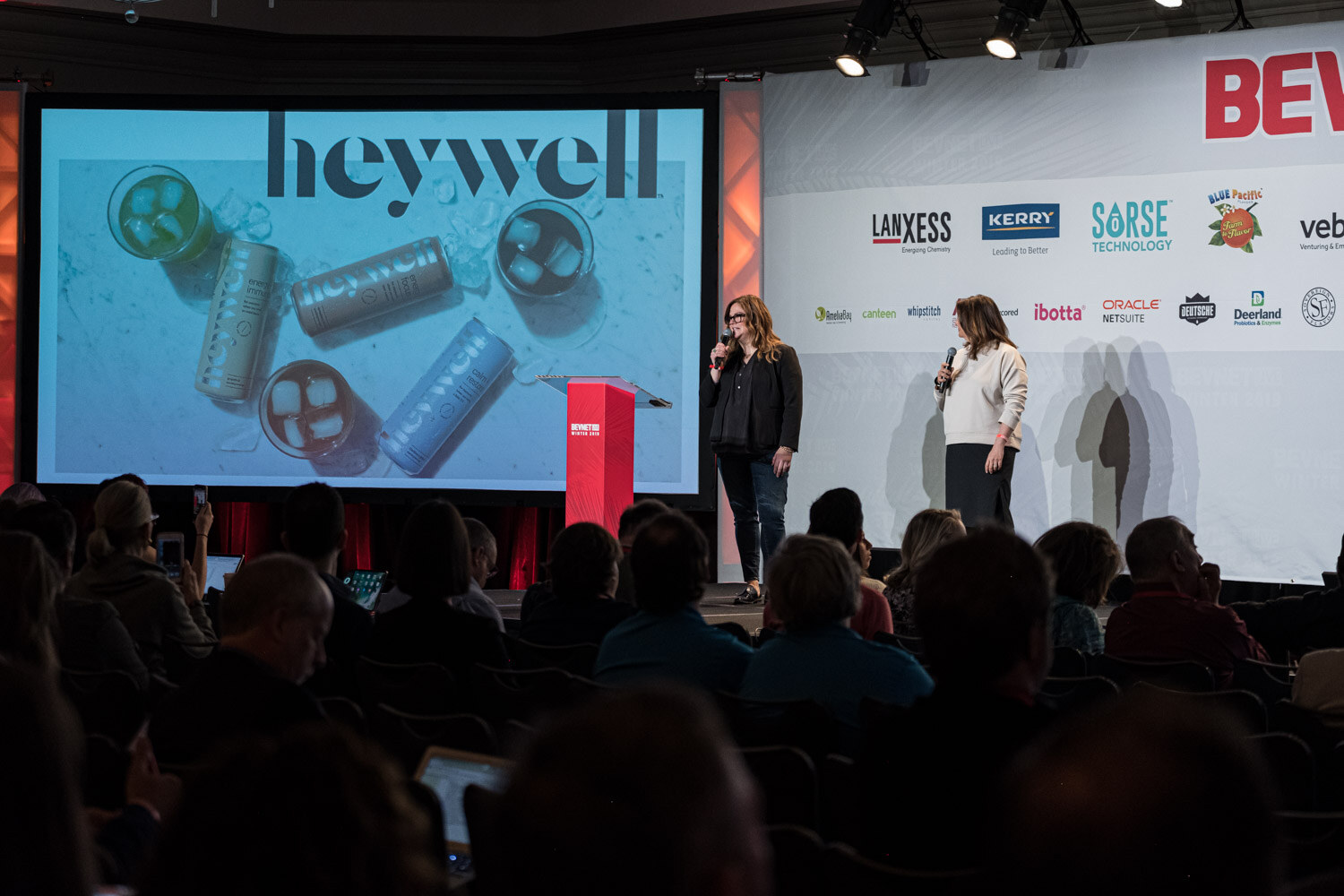 Two women stand at the lectern in front of a large screen with the heywell logo and blue cans in water.
