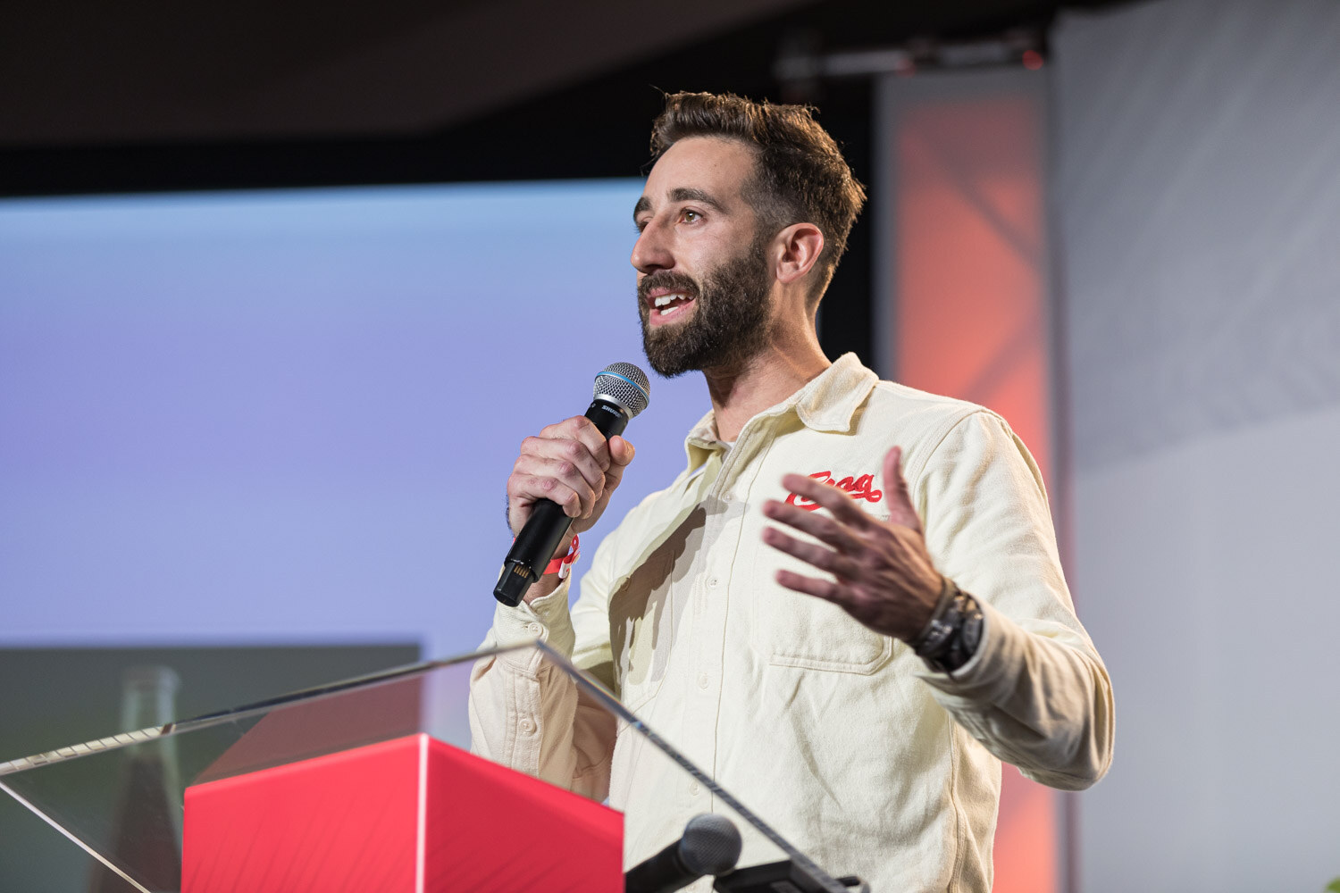 A bearded man in a cream shirt with a red logo holds a microphone and gestures beside the lectern.