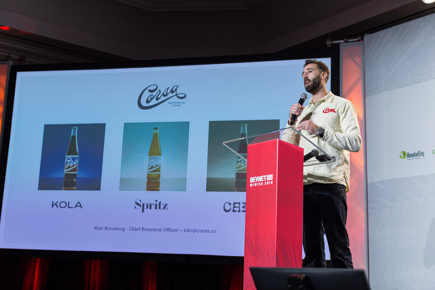 A man in a cream jacket holds a microphone beside a lectern and a screen showing the Corsa logo and three bottles labeled Kola and Spritz.