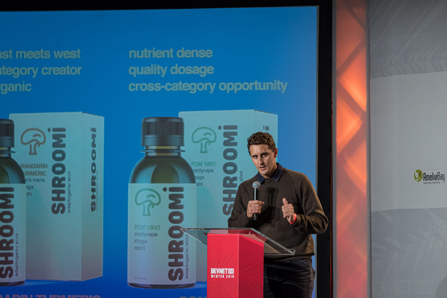 A man in a dark sweater speaks at a lectern in front of a screen showing Shroomi bottles and the words nutrient dense, quality dosage, cross-category opportunity.