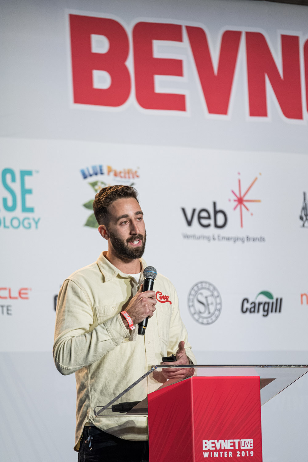 A bearded man in a cream jacket stands at a BevNET Live Winter 2019 lectern with a veb sponsor logo behind him.