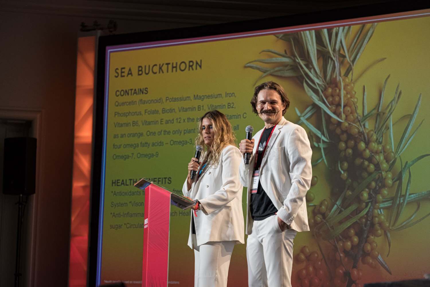 A woman and a man in white outfits hold microphones in front of a large slide titled Sea Buckthorn with photos of the berries.