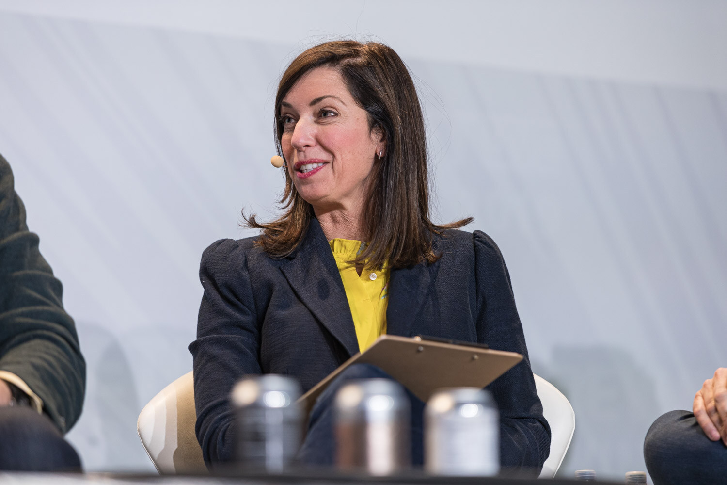 A woman in a navy blazer with a yellow top holds a clipboard with silver cans on the table in front of her.