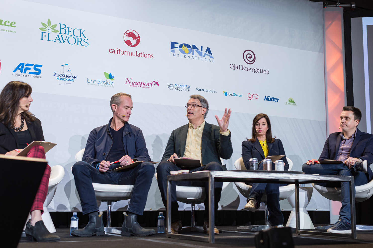 Five people with clipboards sit on white chairs at a low table with silver cans in front of a sponsor wall.