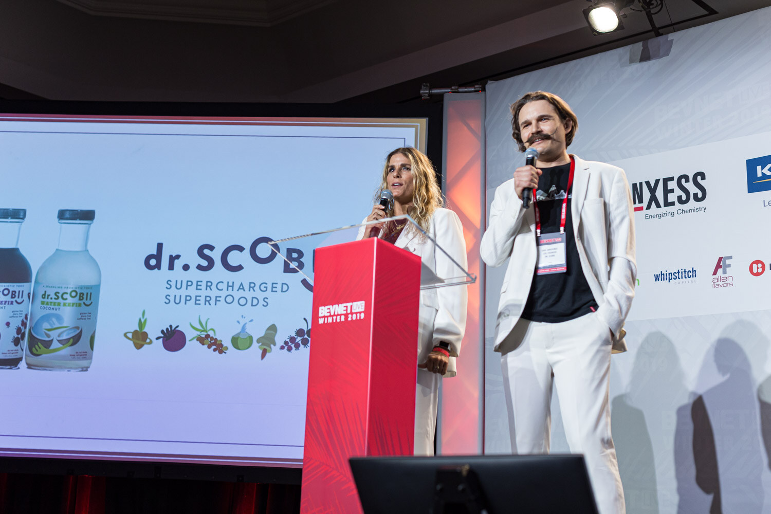 A woman and a man in white outfits stand at a lectern in front of a screen showing dr.Scobi Supercharged Superfoods bottles.