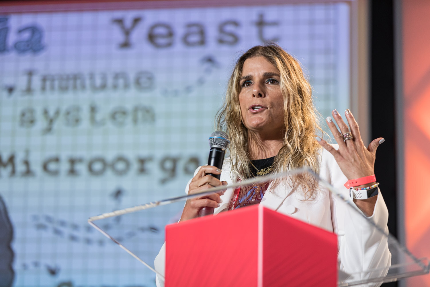A woman with long blonde hair in a white blazer holds a microphone at a clear lectern with a slide reading Yeast, Immune system, Microorganisms.