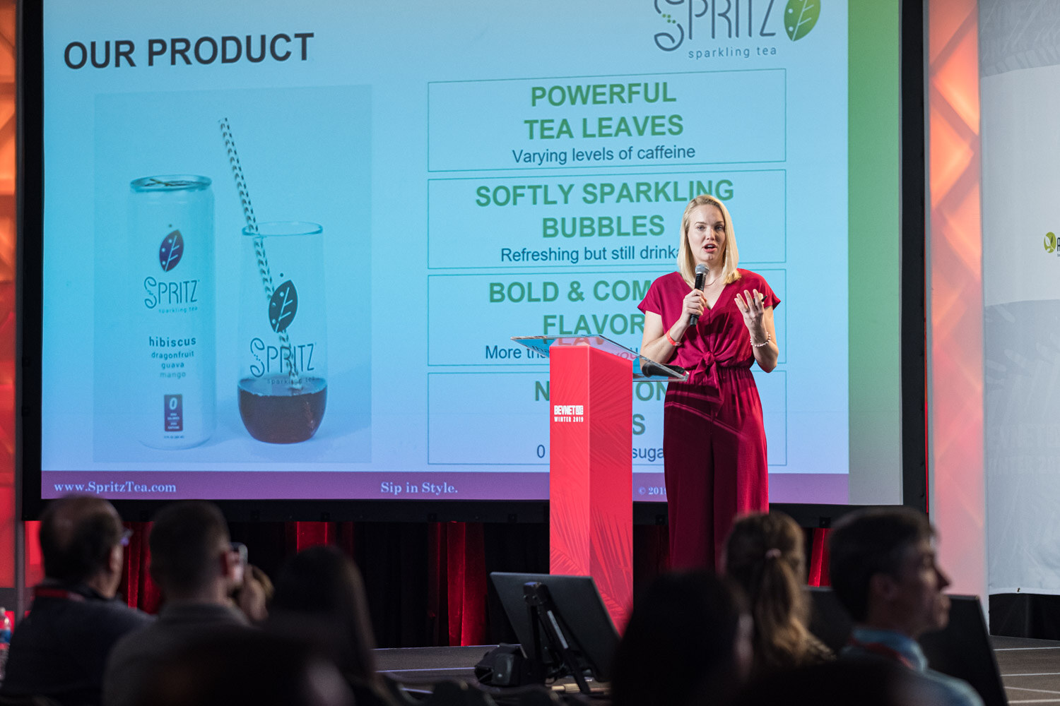 A woman in a red jumpsuit stands at the lectern beside a screen titled Our Product describing Spritz sparkling tea.