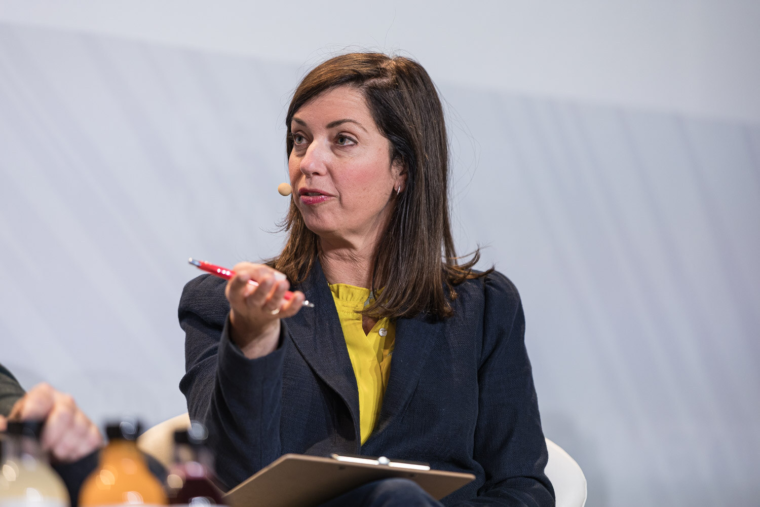 A woman in a navy blazer and yellow top holds a red pen and a clipboard with cans on the table in front of her.