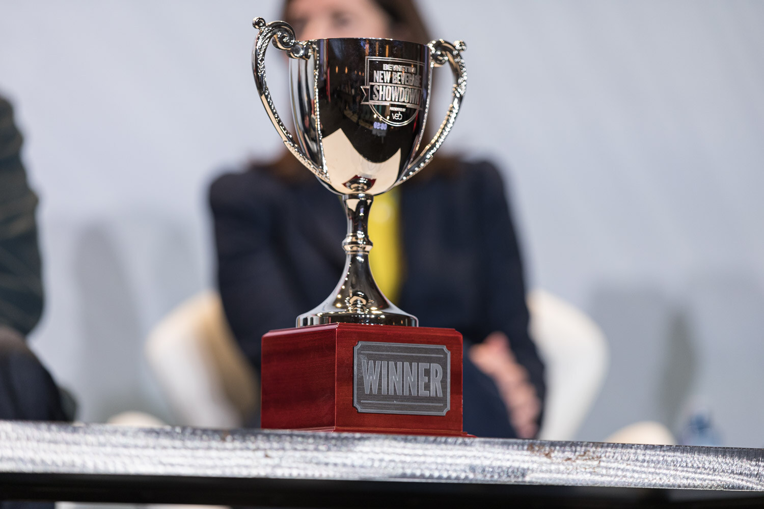 A close view of a silver cup with a plaque reading Winner on a table in front of a blurred seated person.