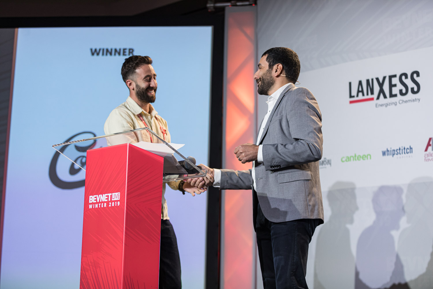Two men shake hands at a red lectern in front of a screen reading Winner with the Corsa logo.