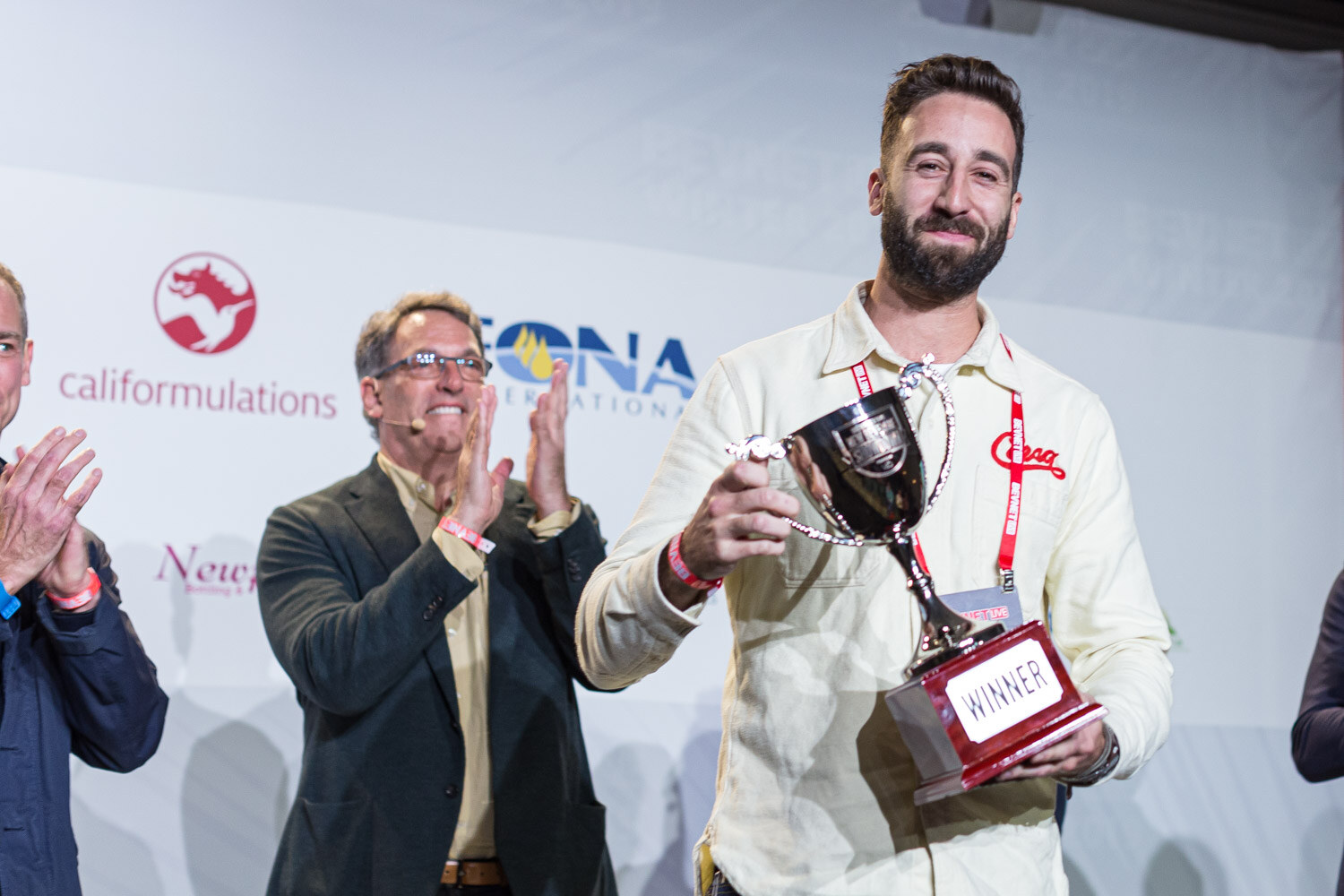 A bearded man in a cream Corsa shirt holds up a silver cup on a base labeled Winner while a man in a dark jacket applauds.
