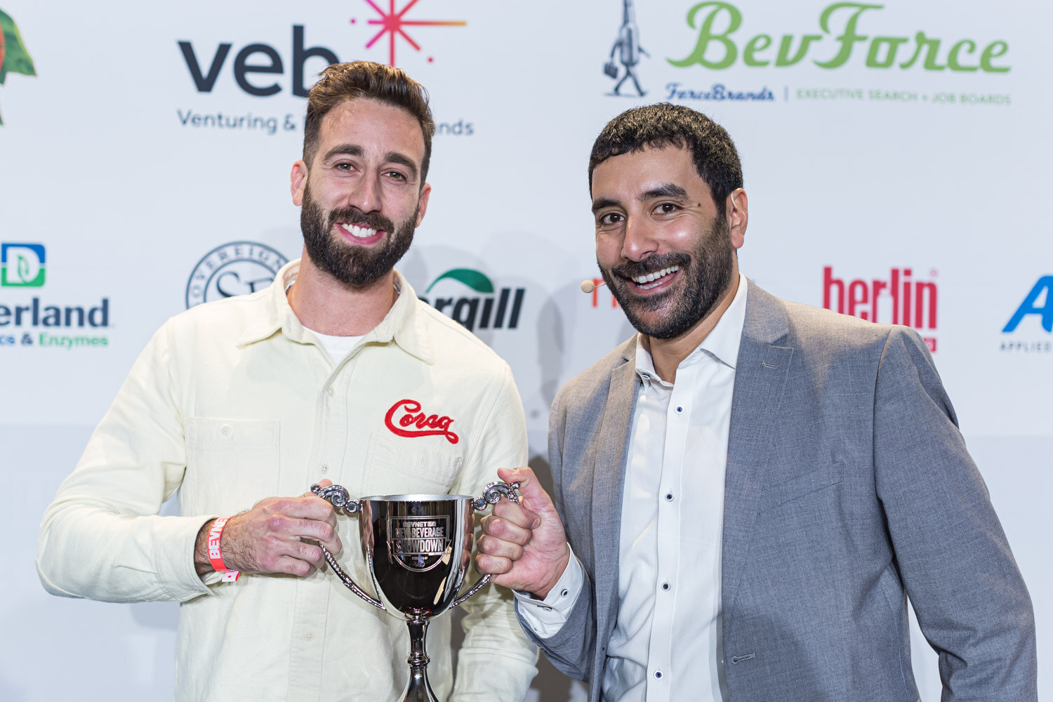 A man in a cream Corsa shirt holds a silver trophy cup while a smiling man in a grey blazer stands beside him in front of the veb and BevForce sponsor wall.