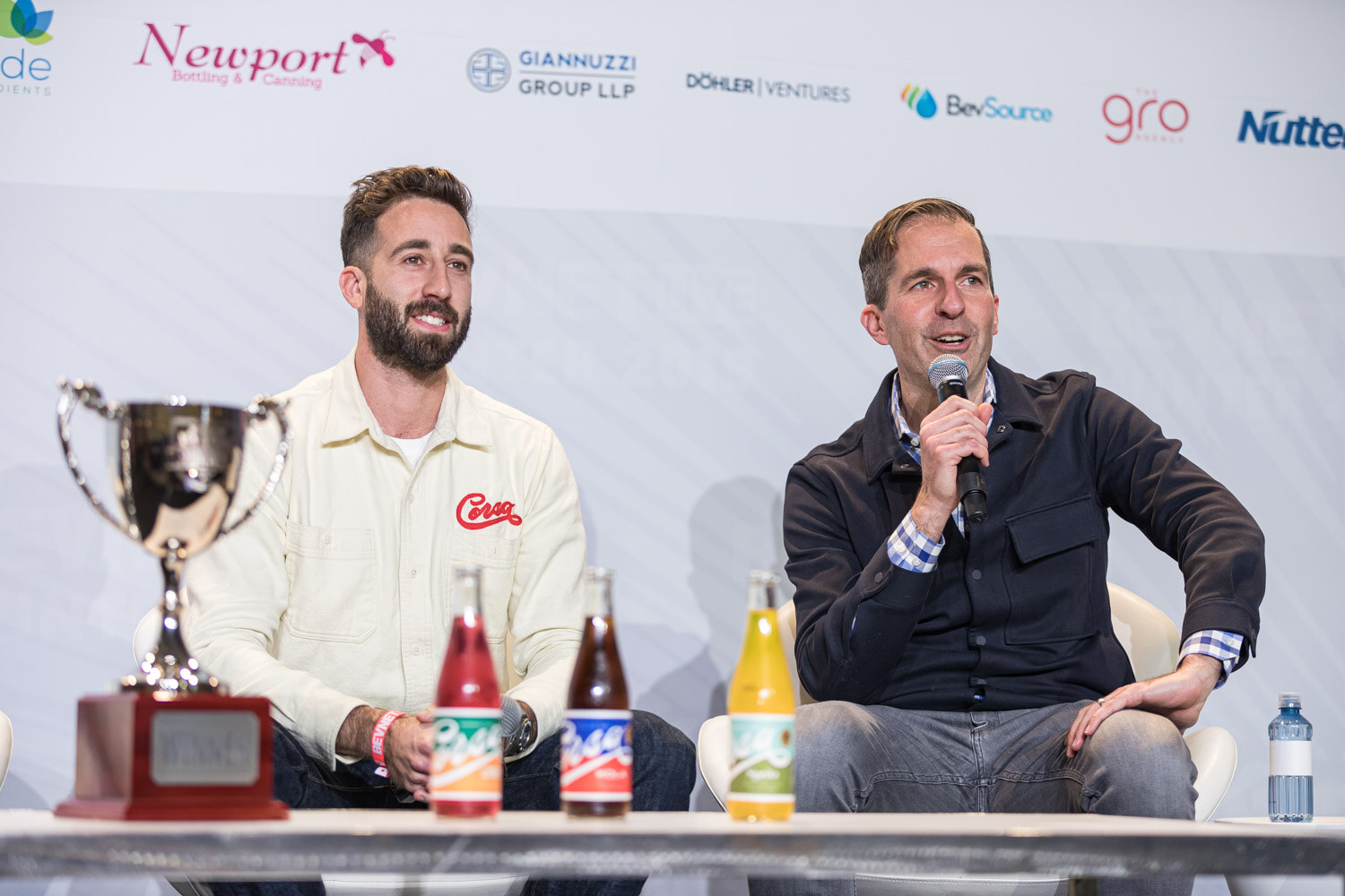 A bearded man in a cream Corsa shirt and a man in a dark jacket holding a microphone sit beside a silver trophy and three Corsa bottles.