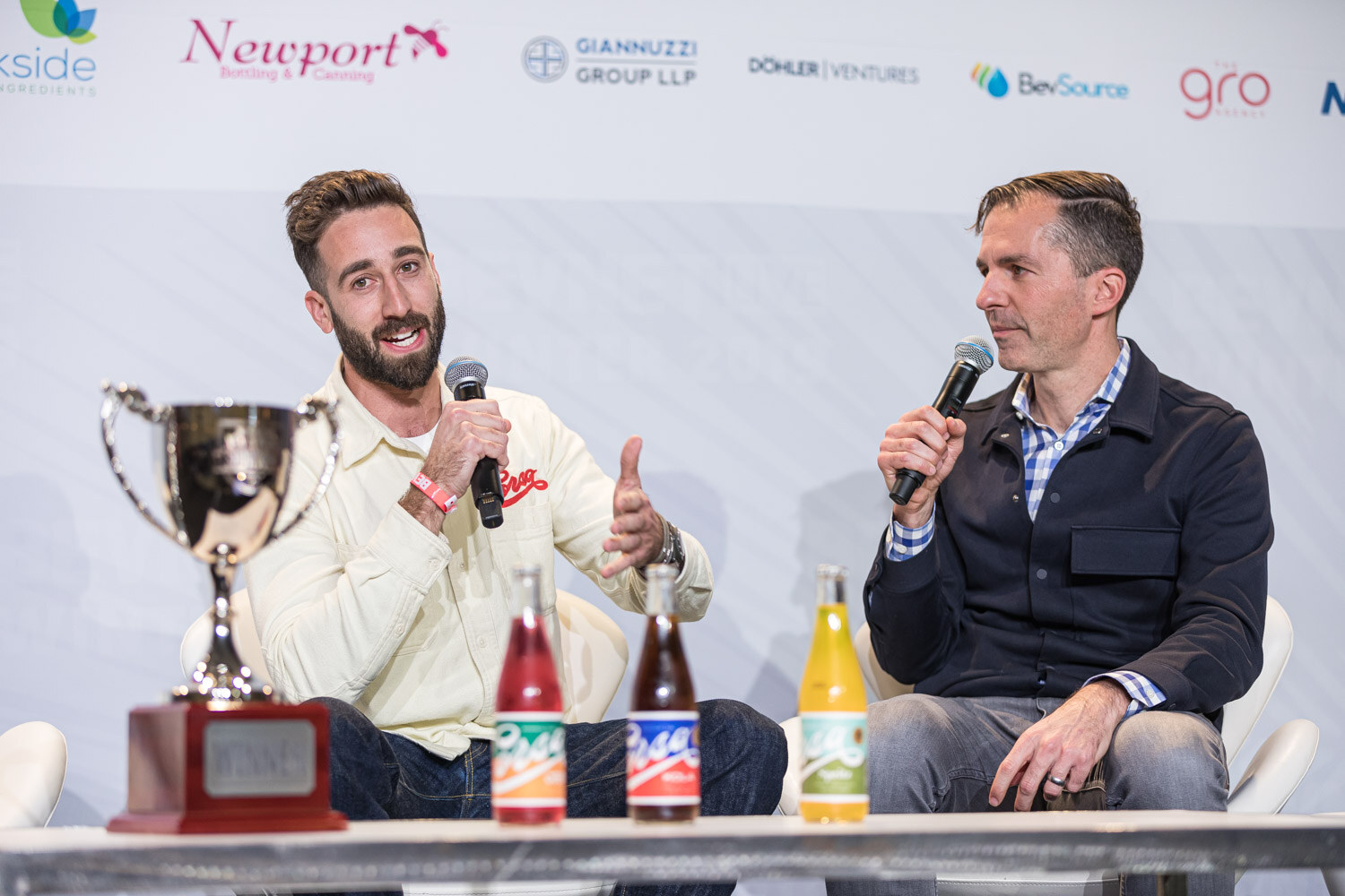 A man in a cream Corsa shirt gestures with a microphone beside a silver trophy and Corsa bottles as a man in a navy jacket listens.