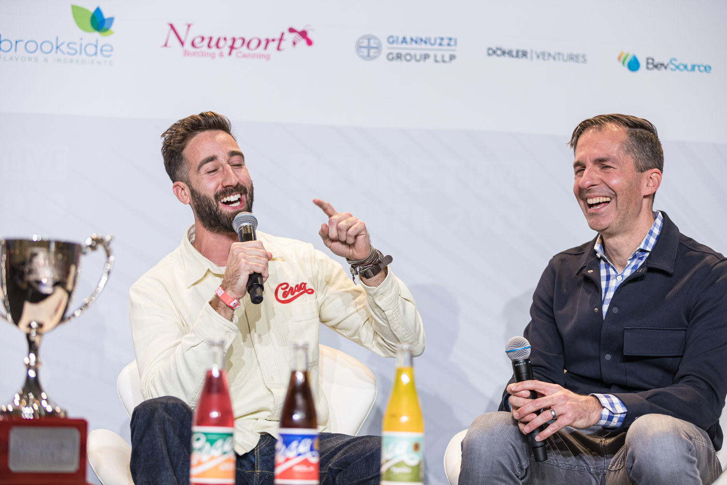 A man in a cream Corsa shirt points while laughing next to a man in a navy jacket who is also laughing, with a trophy and bottles on the table.