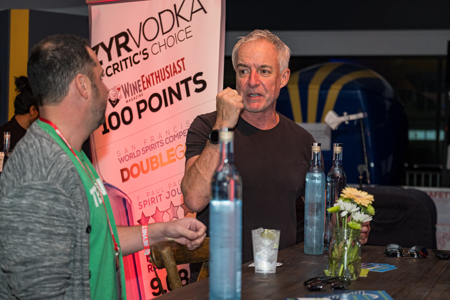 Two men stand at a table with water, glasses and bottles in front of a vodka sign advertising 100 points.