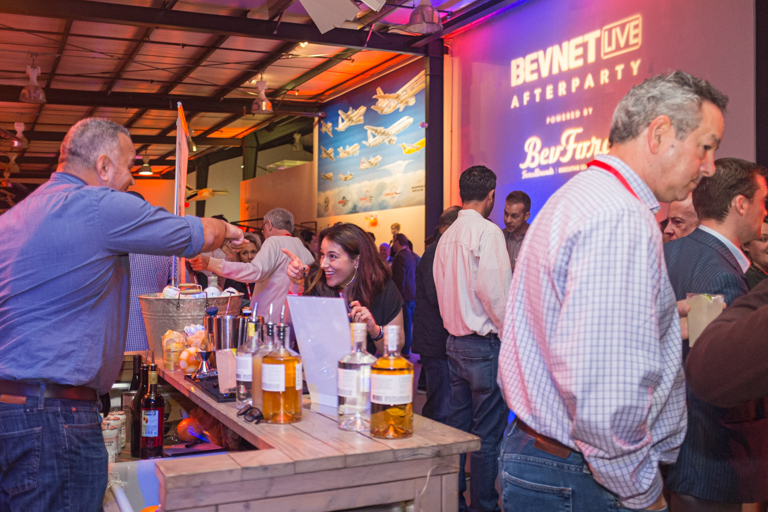 Guests stand at a bar with whiskey bottles under a lit BevNET Live Afterparty sign inside an aircraft museum hangar.