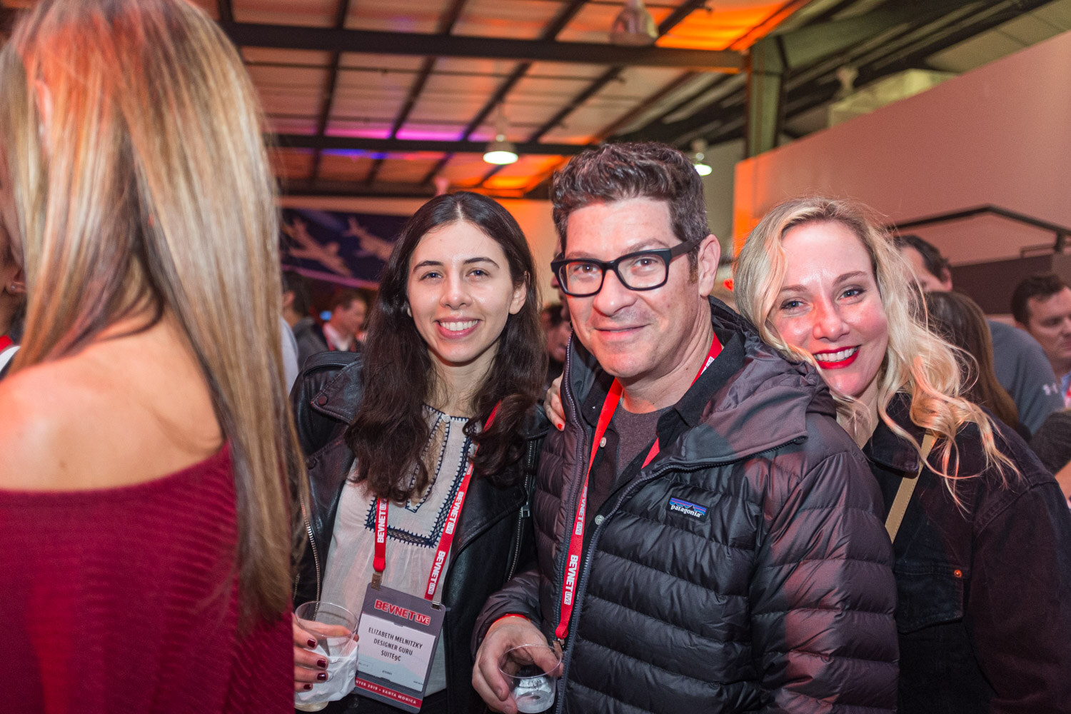 Two women and a man in glasses wearing red lanyards smile close together inside a dimly lit museum hall.