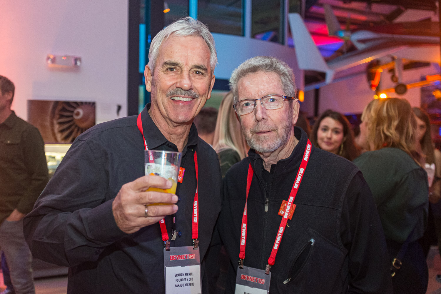 Two older men with red lanyards pose side by side, one holding a glass of beer, with museum exhibits behind them.