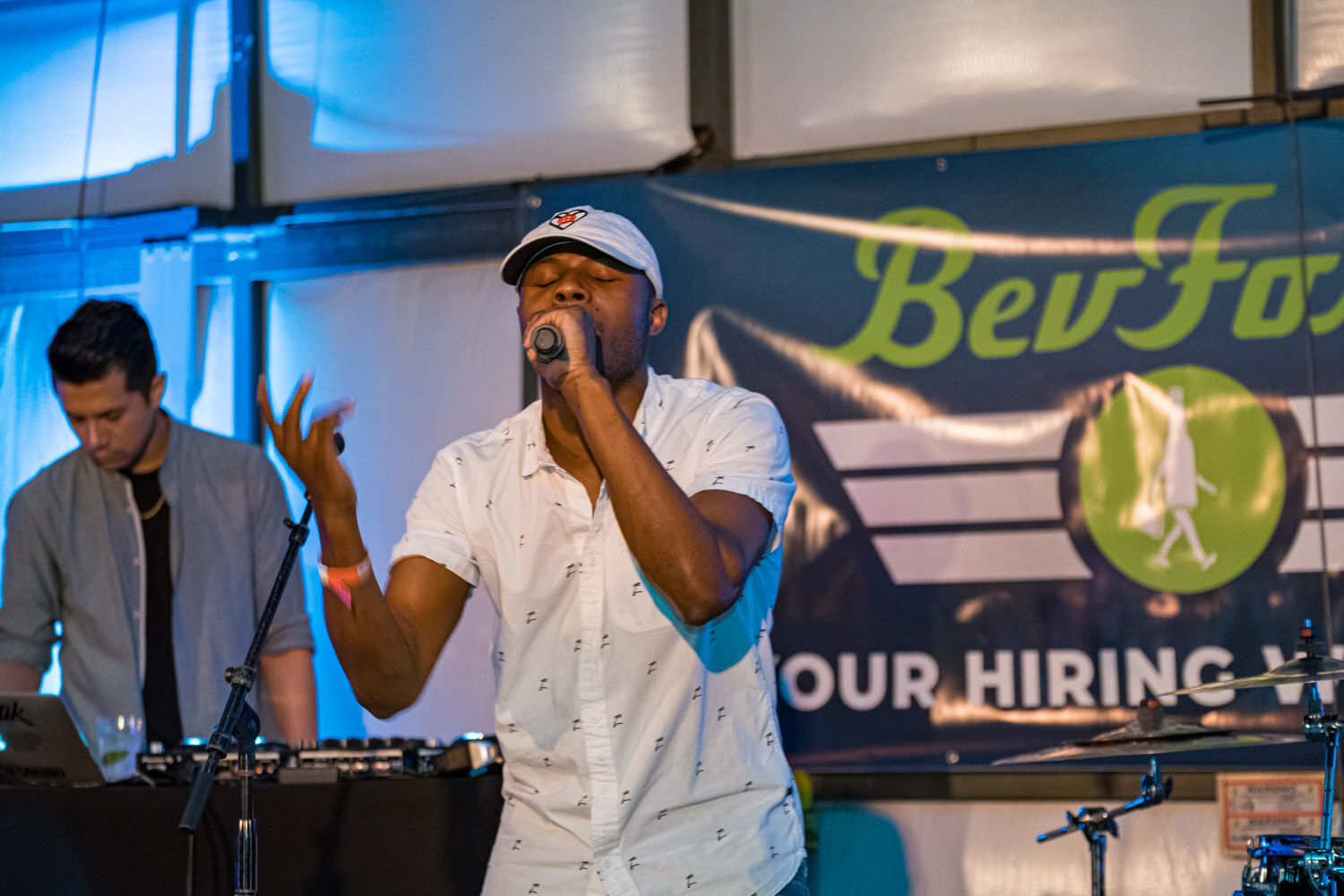 A singer in a white shirt and cap holds a microphone in front of a BevForce banner while a DJ works behind him.