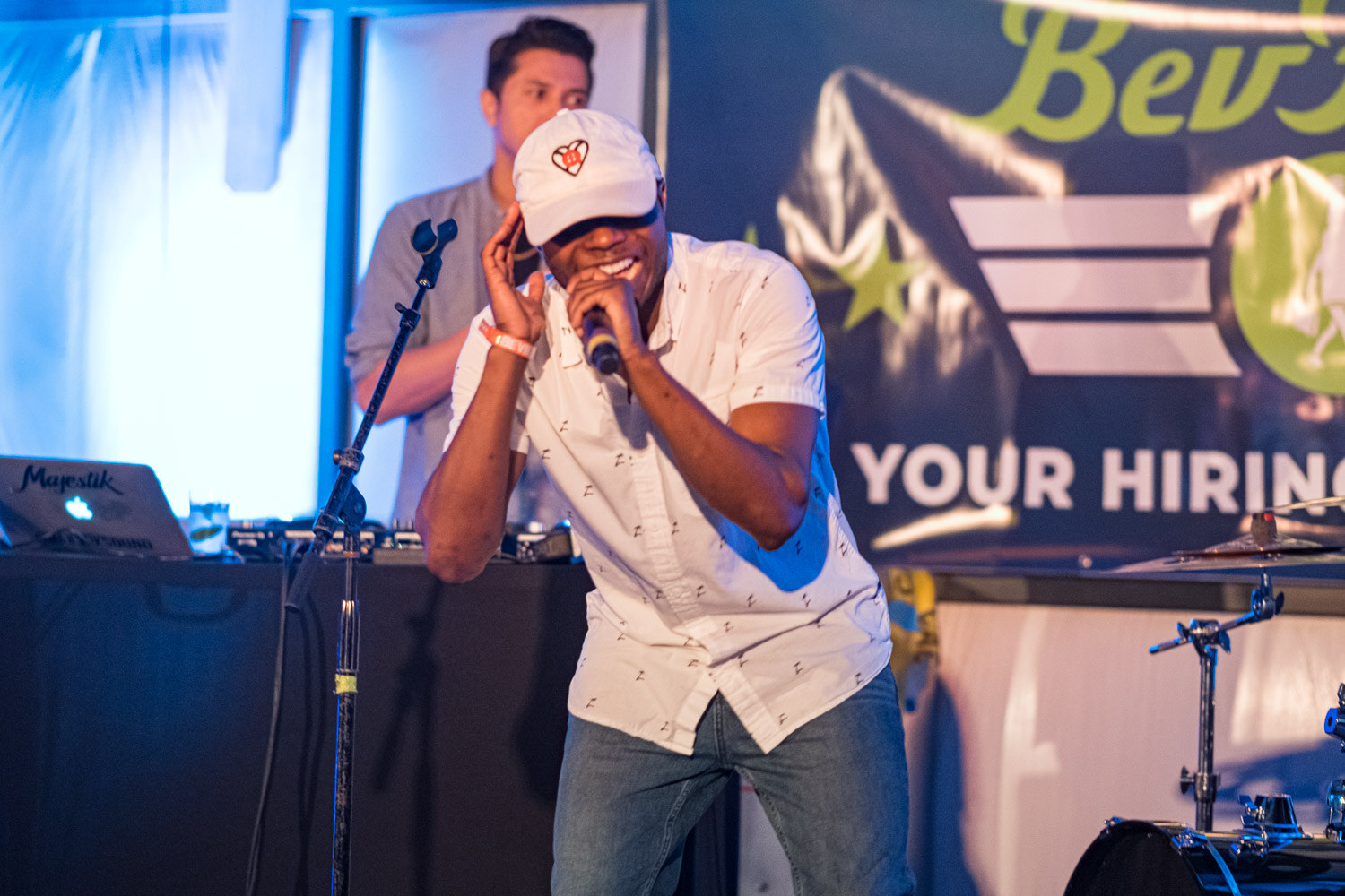 A man in a white cap and white patterned shirt sings into a microphone in front of a DJ setup and a BevForce banner.