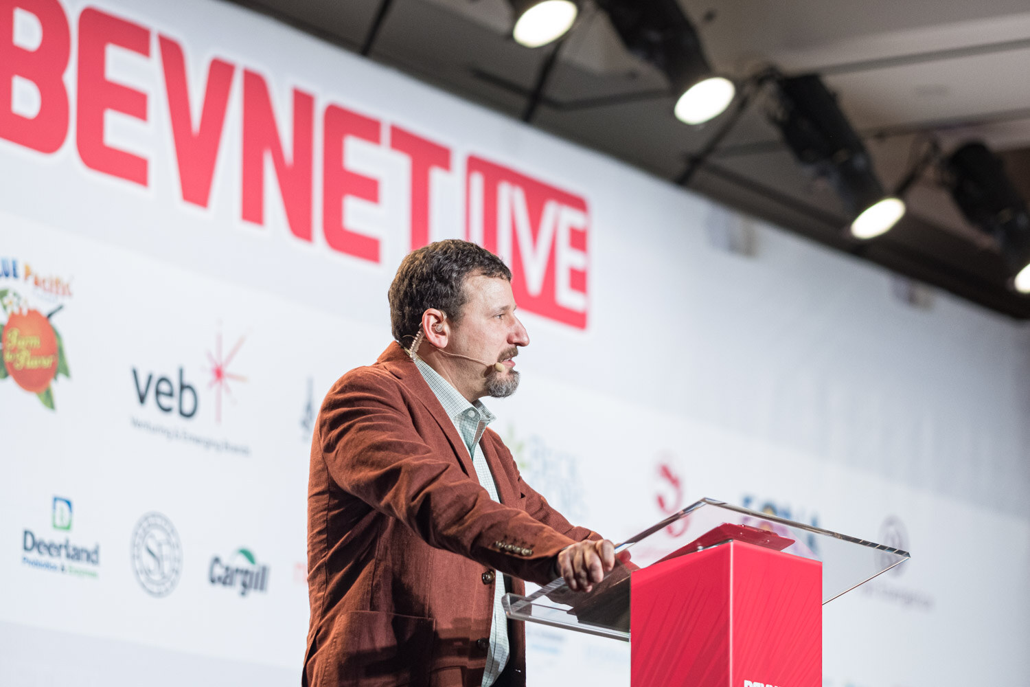 A bearded man in a rust jacket speaks from a red lectern in front of a BevNET Live sponsor wall.