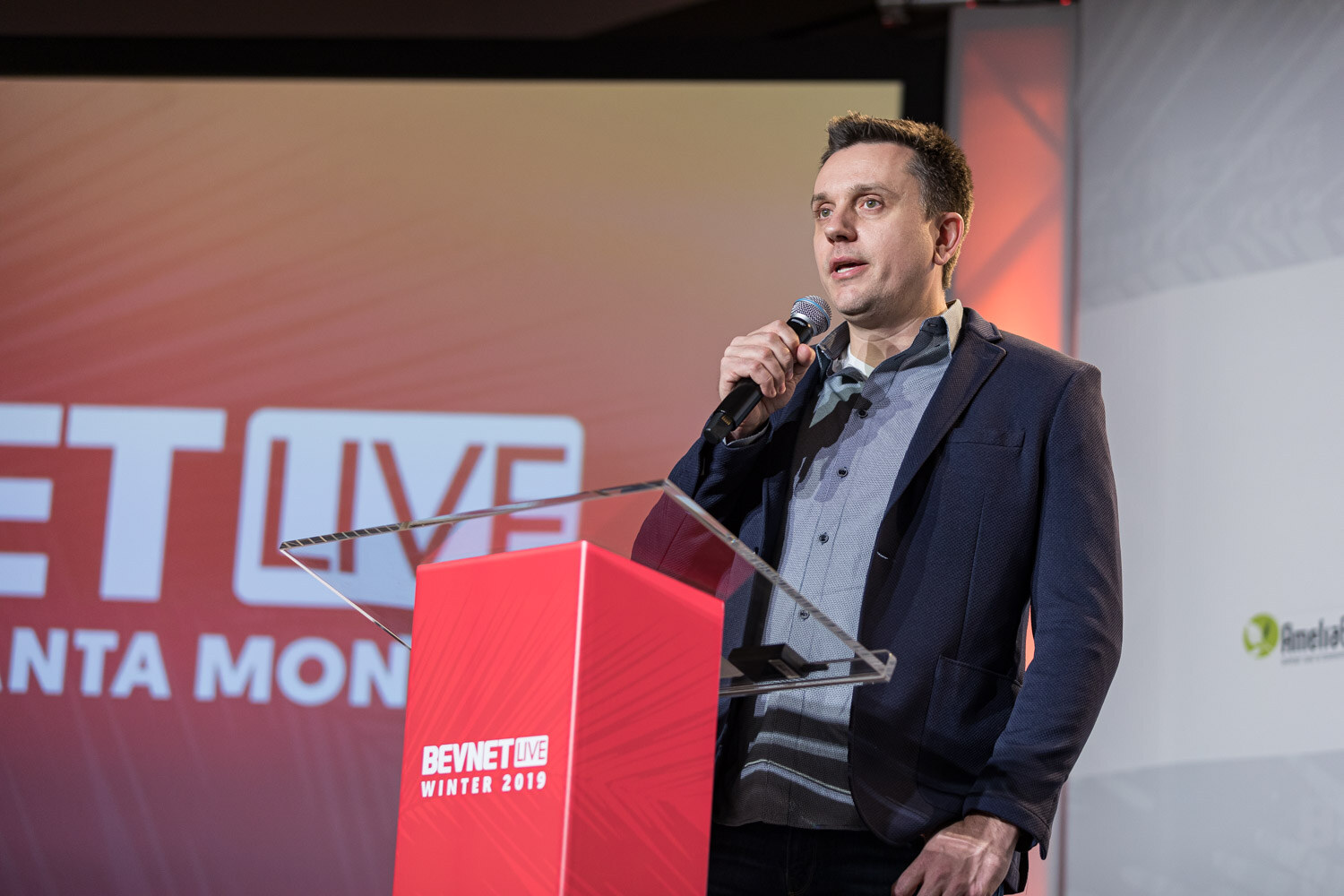 A man in a navy blazer holds a microphone beside a red lectern in front of a magenta BevNET Live screen.