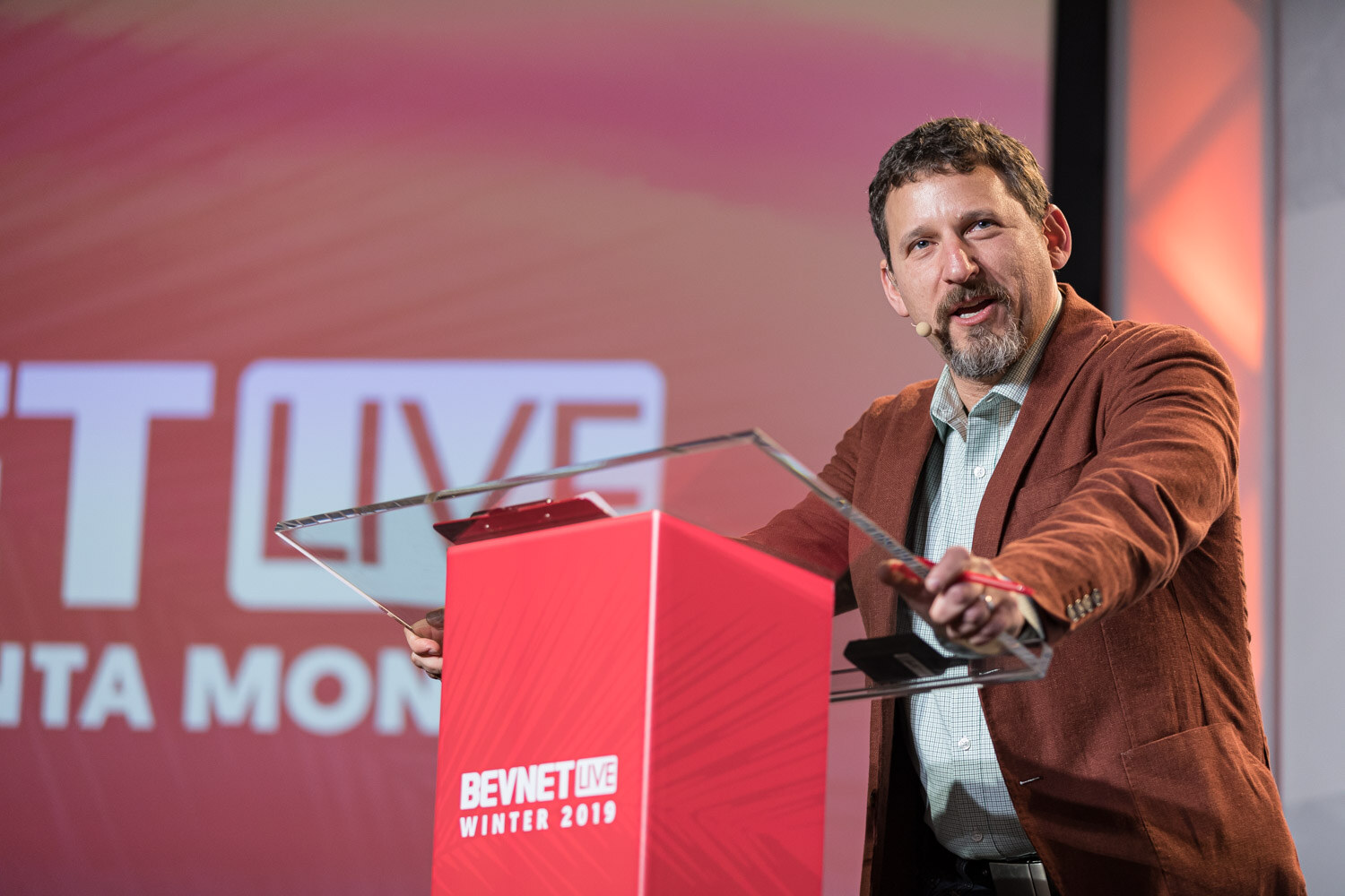 A bearded man in a rust jacket gestures at a clear lectern in front of the BevNET Live Winter 2019 screen.