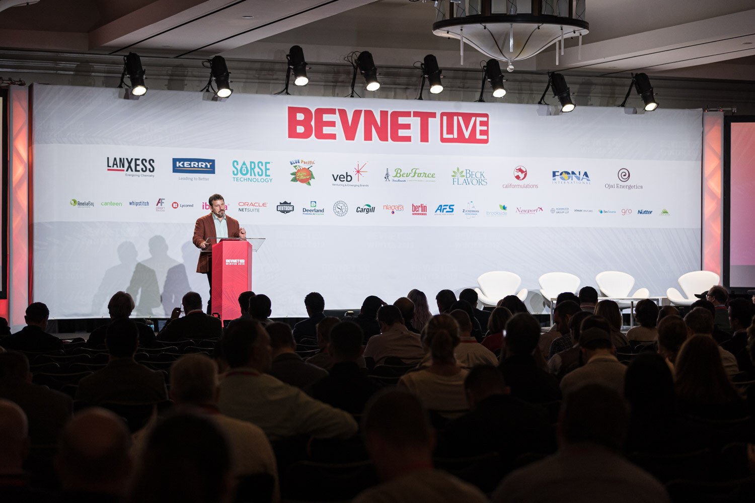 A wide view of a ballroom with a speaker at a red lectern on stage and rows of attendees in silhouette.