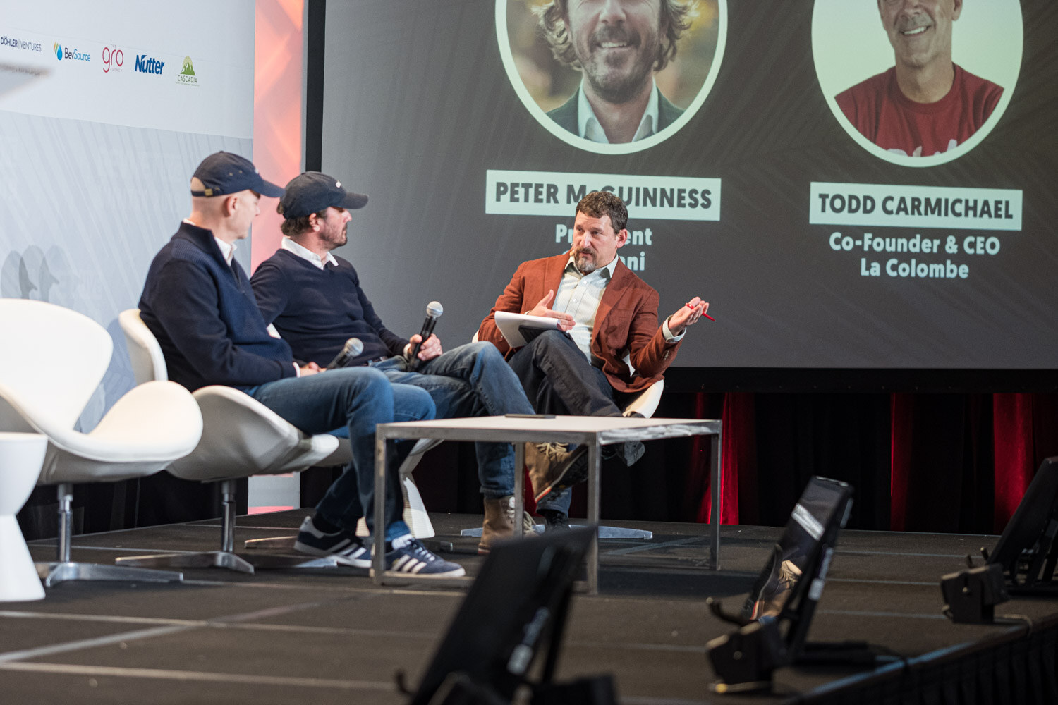 Two men in dark sweaters sit on white chairs with a moderator in a rust jacket in front of a screen showing Peter McGuinness and Todd Carmichael.