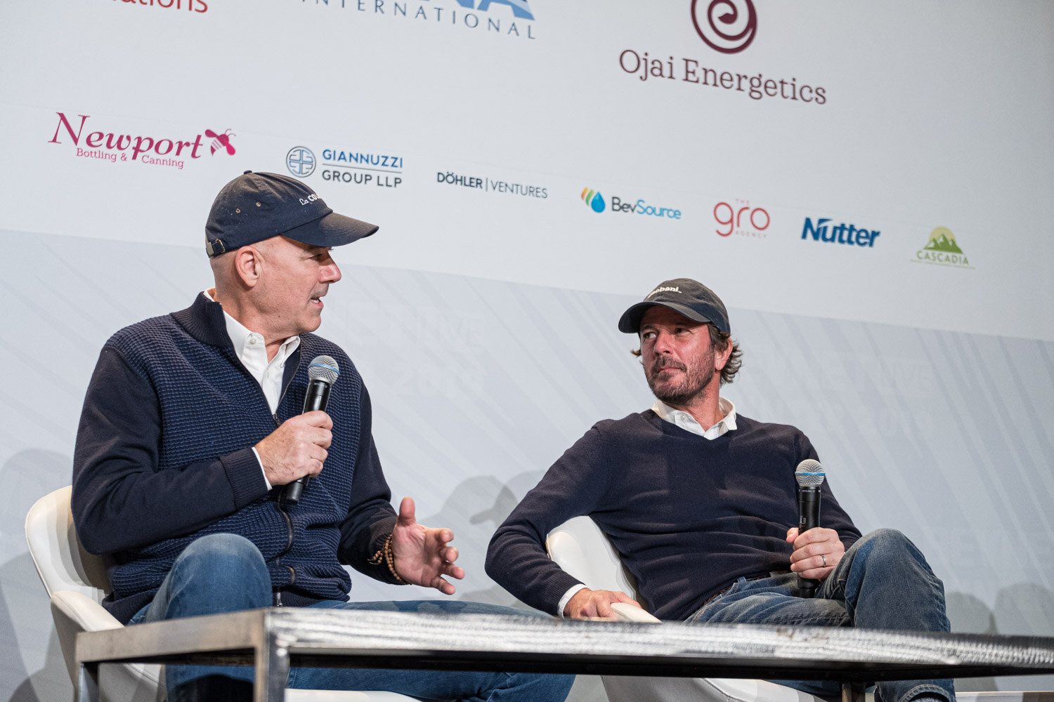 Two men in navy sweaters and baseball caps sit with microphones in front of a sponsor wall.