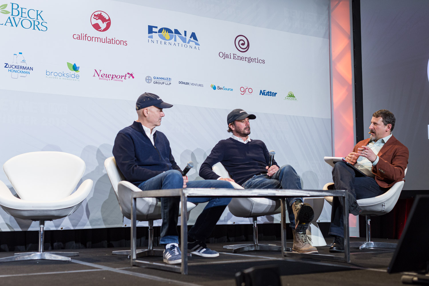 A wide shot of a stage with three people in white swivel chairs and a table in front of a sponsor wall.