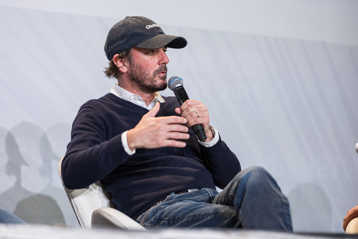 A bearded man in a baseball cap and navy sweater gestures with a microphone in hand.