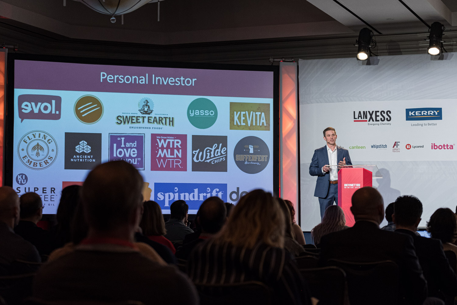 A man in a navy suit stands at a lectern beside a screen showing logos such as Sweet Earth, Kevita, I and love and you, and Wilde under the title Personal Investor.